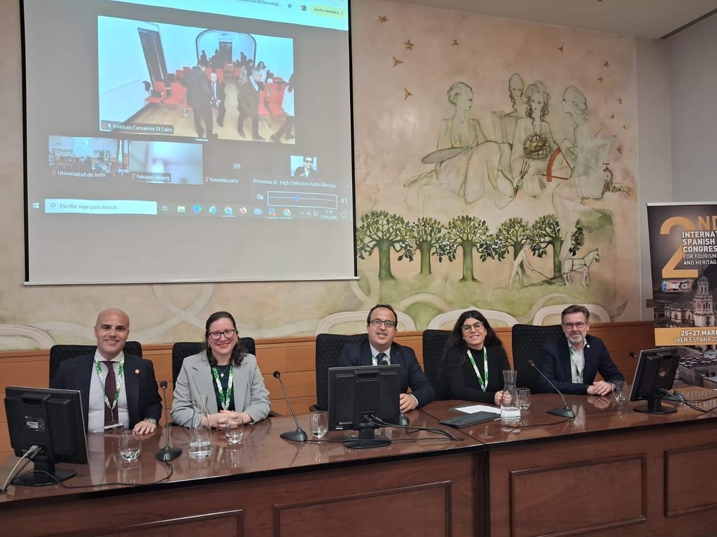 Five panellists sit along a long conference table beneath a screen carrying a live video link with a partner institution.