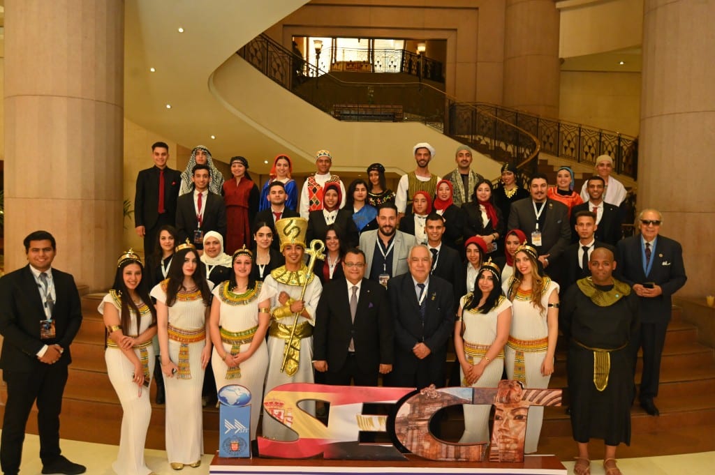 A large group poses on a grand staircase, with a man in a gold pharaoh costume holding an ankh staff at the centre front row.