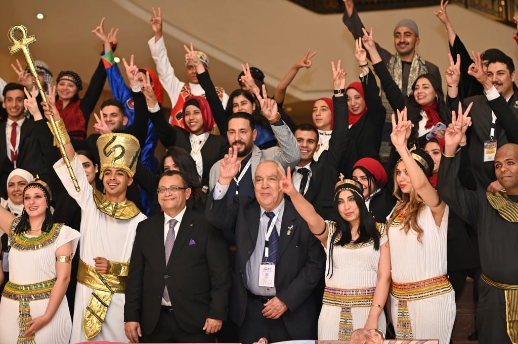 A cheering crowd of delegates and hosts in pharaonic costume raise their hands together in celebration on a staircase.