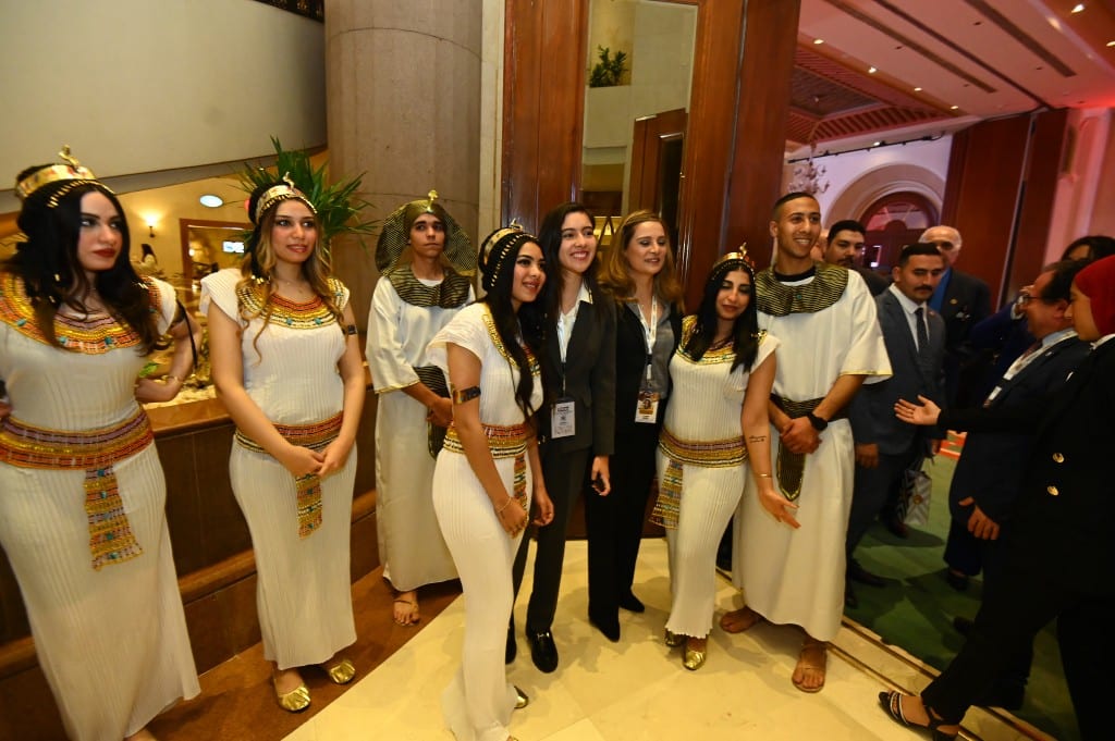 Women in white and gold pharaonic costume with beaded headdresses greet a visitor at a hotel entrance.