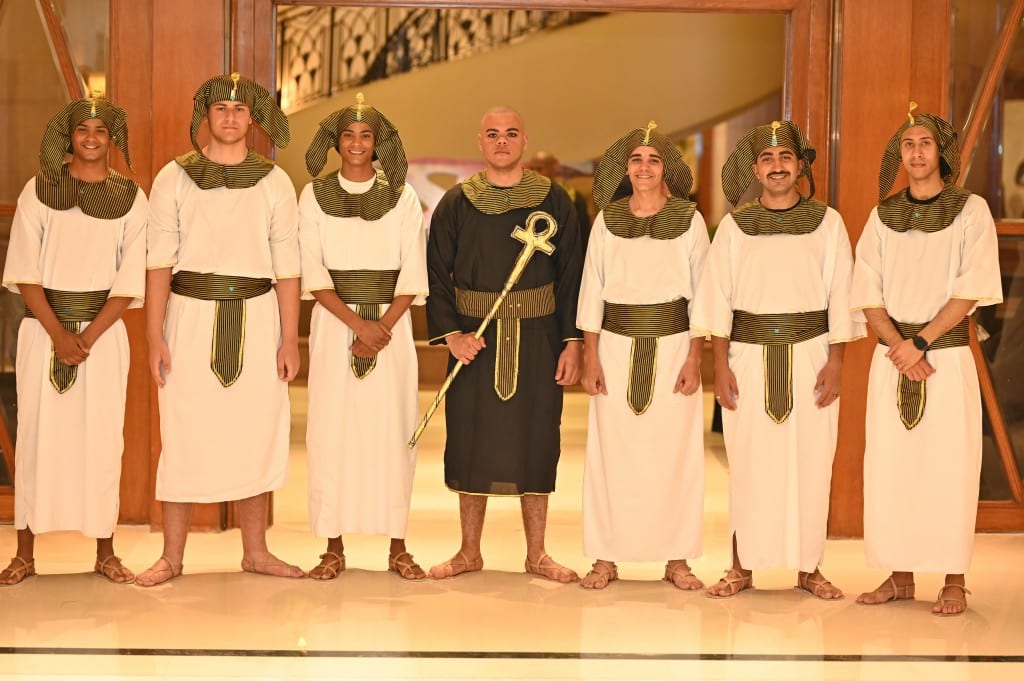Seven hosts in matching white and gold pharaonic robes and headdresses stand in a row, one holding a gold ankh.
